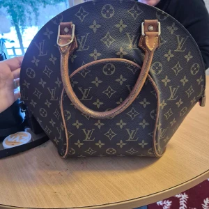 Brun handväska från Louis Vuitton - Säljer en elegant brun handväska från Louis Vuitton med det klassiska monogrammönstret. Väskan har rund form och två handtag i skin, lite sliten i handtagen se bild. Äkthetsintyg saknas men går att se äkthet på serienummer 