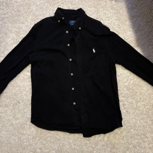 Ralph lauren skjorta  - Svart Ralph lauren skjorta med vit logga på. Skjortan är i väldigt bra skick och de finns inga tecken på användning. Den är i ett mjukt material. 