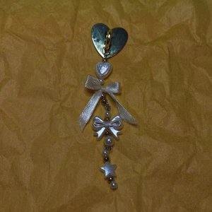 MOBILSMYCKE - WHITE BOW & PEARLS - 🫶🏼handgjorda accessoarer🫶🏼 ✨I varje köp ingår klistermärken✨ 💛 Mobilsmycket har en klistrig-yta på baksidan som man enkelt kan sätta fast på mobilskalet eller andra platta ytor 💛  ”Charmigt hängsmycke med ett stort guldfärgat hjärta, pärlor och en stjärna. Smycket har en söt rosett och flera små detaljer som ger en elegant touch. Perfekt för att dekorera en väska eller mobil.”