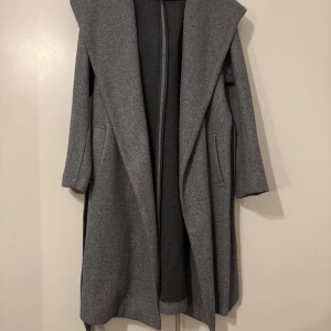 Grå kappa från Zara - Snygg grå kappa från Zara med huva och knytbälte i midjan. Perfekt för kyligare dagar med sin långa design och praktiska fickor. En stilren och mångsidig jacka för alla tillfällen. Jackan är köpt för 550kr knappt använd och även kemtvättad utan användning efter. Kan fraktas men köparen står för frakten :) 