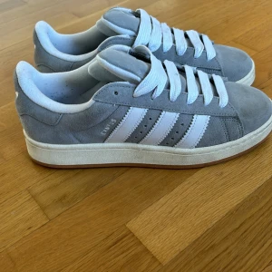 Grå Adidas Campus sneakers - Säljer ett par snygga grå Adidas Campus sneakers i mocka. De har de klassiska tre vita ränderna på sidan och en vit sula. Skorna har snörning och är i bra skick.  Perfekta för både vardag och fest! Jag har använt de endast 1 gång