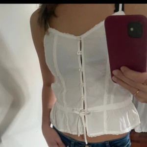 Vit topp från Brandy Melville - Säljer en superfin vit topp från Brandy Melville. Den har smala axelband och knappar framtill, vilket ger en söt look. Toppen är croppad med en liten volangkant längst ner. Den går att dra åt i midja vilket gör att man kan välja om man vill ha den tajt eller lite lösare. Prislappen är kvar då den aldrig är använd, köpt i Barcelona. Är One size men är som en Xs, S. Nypris 286 kr, säljer för 120🤗💕