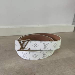 Säljer ett snyggt vitt bälte från Louis Vuitton med deras klassiska monogrammönster. Bältet har en stor LV-spänne i silverfärg och är gjort av skinn. Perfekt för att ge din outfit en lyxig touch!