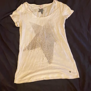 Vit t-shirt med stjärna från Esprit - Säljer en snygg vit t-shirt från Esprit med en stor glittrig stjärna på framsidan. T-shirten är kortärmad och har en rund halsringning. Perfekt för en avslappnad stil eller att glamma upp en vardagsoutfit. Den är i perfekt skick och redo för nya äventyr! 