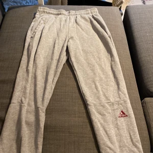 Grå mjukisbyxor från Adidas - Säljer ett par grå mjukisbyxor från Adidas. De är superbekväma och perfekta för chill eller träning. Byxorna har en elastisk midja och det klassiska Adidas-loggan i rött på benet. Perfekta för en avslappnad stil!