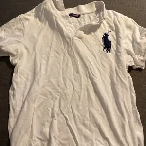 Säljer en klassisk vit t-shirt från Ralph Lauren med korta ärmar. Den har en stor, broderad logga i mörkblått på bröstet. Perfekt för en avslappnad och stilren look. T-shirten är i bomull och passar bra till både vardag och fest. Skrynklig för att inte använt på länge men en tvätt och strykjärnet löser det