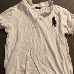 Vit kortärmad t-shirt från Ralph Lauren - Säljer en klassisk vit t-shirt från Ralph Lauren med korta ärmar. Den har en stor, broderad logga i mörkblått på bröstet. Perfekt för en avslappnad och stilren look. T-shirten är i bomull och passar bra till både vardag och fest. Skrynklig för att inte använt på länge men en tvätt och strykjärnet löser det