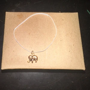 Armband med elefantberlock - Säljer ett enkelt och stilrent armband med en liten elefantberlock i guld. Armbandet har en tunn vit tråd som ger en minimalistisk look. Perfekt för vardagsbruk eller som en söt present!