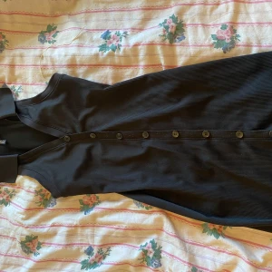 Svart klänning - Säljer en elegant svart ärmlös klänning i ribbat material. Klänningen har en stilren design med knappar längs framsidan och en klassisk krage. Perfekt för både vardag och festliga tillfällen! Pris kan diskuteras 💗