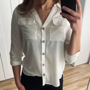 Vit blus med knappar - Stilren vit blus med knappar framtill. Blusen har långa ärmar som kan rullas upp och fästas med knapp. Har några små fläckar på insidan, men syns inte när man har den på sig.