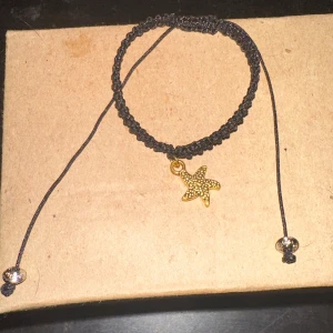 Svart armband med sjöstjärna - Säljer ett snyggt svart armband med en guldig sjöstjärna som hänge. Armbandet är flätat och justerbart med två små silverfärgade pärlor i ändarna. Perfekt för att ge en strandig känsla till din outfit!