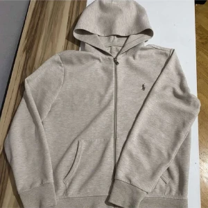 Polo ralph lauren Zipup - Säljer en stilren beige hoodie från Polo Ralph Lauren med dragkedja och klassisk logga på bröstet. Perfekt för en avslappnad look. Den har långa ärmar och en praktisk huva.