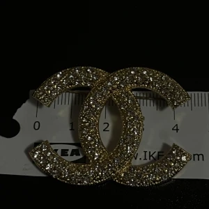Guldglittrig brosch - Elegant brosch i form av två sammanflätade C:n, täckt med gnistrande stenar. Perfekt för att ge en lyxig touch till din outfit. Helt ny! Storlek  syns på bilderna ✨☺️