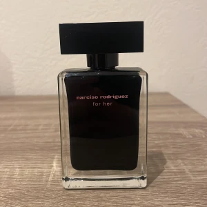 Narciso Rodriguez For Her Eau de Toilette - Elegant parfymflaska från Narciso Rodriguez med en stilren svart design. Flaskan är rektangulär med en svart kork och rymmer 50 ml av doften 'For Her'. Perfekt för den som söker en sofistikerad och tidlös doft.