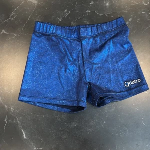 Blå glittriga shorts från Quatro - Snygga blå glittriga shorts från Quatro, perfekta för att sticka ut. De har en elastisk midja och är tillverkade i en blandning av nylon och spandex för en bekväm passform. Den används främst för att ha över sina gymnastik dräkter men kan anvöndas som vanliga tränings short också 