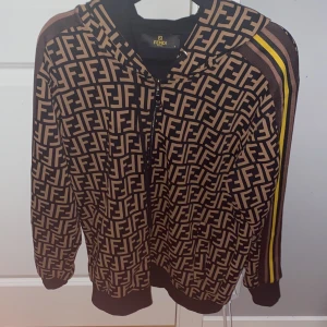 Mönstrad hoodie från Fendi - Säljer en snygg hoodie från Fendi med det ikoniska FF-mönstret i svart och beige. Tröjan har en dragkedja framtill och randiga detaljer i gult och brunt längs ärmarna. Perfekt för dig som vill ha en stilren och lyxig look.