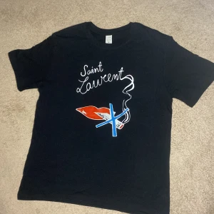 Svart t-shirt med Saint Laurent-tryck - Snygg svart t-shirt med ett coolt tryck från Saint Laurent. Designen har en röd läpp och en blå cigarett med rök, vilket ger en edgy look. Perfekt för dig som vill sticka ut med stil.
