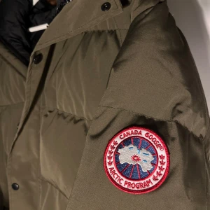 Olivgrön dunjacka från Canada Goose - Säljer en olivgrön dunjacka från Canada Goose med pälskantad huva och broderad logga på ärmen. Jackan har dragkedja och knappar framtill samt flera praktiska fickor. Perfekt för kalla vinterdagar. Har använts 2 gånger