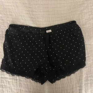 Svarta pyjamasshorts med spetsdetalj - Snygga svarta shorts med vita prickar och spetsdetaljer vid bensluten. De har en bekväm passform med knytband i midjan. Perfekta för en avslappnad stil.