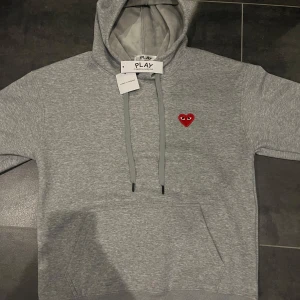 Grå hoodie från Comme des Garçons - Säljer en stilren grå hoodie från Comme des Garçons med det ikoniska röda hjärtat på bröstet. Tröjan har en justerbar huva med dragsko och en praktisk magficka. Perfekt för en avslappnad look. 