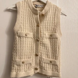 Beige stickad väst från Zara - Snygg beige stickad väst från Zara med ett klassiskt flätmönster. Västen har knappar framtill och två praktiska fickor. Perfekt för lager-på-lager-stil.
