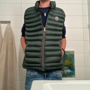 Monclee väst - Pris går att diskutera vid snabb affär. Snygg grön dunväst från Moncler med dragkedja och logotyp på bröstet. Perfekt för lager-på-lager under kyliga dagar. Västen har en stilren design med quiltade paneler.  Modellen är 175cm lång 