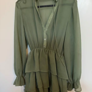 Grön klänning med volanger - Snygg grön klänning med volanger och v-ringning. Den har långa ärmar med knappar framtill och en luftig passform. Perfekt för en stilren look.