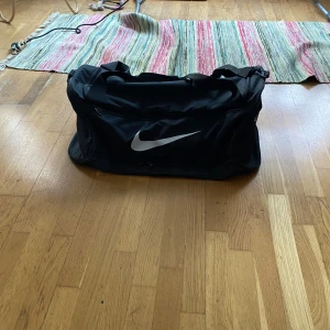 Svart sportväska från Nike - Säljer en svart sportväska från Nike med det klassiska swoosh-loggan i vitt. Väskan har flera fack med dragkedja och justerbar axelrem för bekväm bärning. Perfekt för träning eller resor.