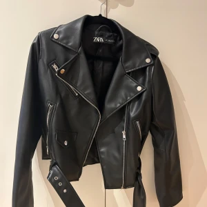 Svart skinnjacka från Zara - Snygg svart skinnjacka från Zara med coola dragkedjedetaljer och knappar. Jackan är knappt använd och i super fint skick!