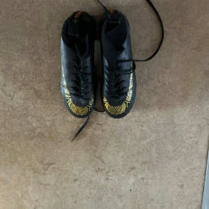 Svarta fotbollsskor med gulddetaljer - Snygga svarta fotbollsskor med gulddetaljer och orange rand på hälen. Skorna har snörning och är perfekta för fotbollsplanen.