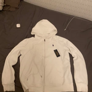 Vit hoodie från Polo Ralph Lauren - Säljer en stilren vit hoodie från Polo Ralph Lauren med dragkedja och den klassiska loggan broderad på bröstet. Perfekt för en avslappnad look. Hoodien har långa ärmar och en bekväm passform.
