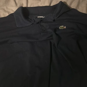 Mörkblå pikétröja från Lacoste - Säljer en klassisk mörkblå pikétröja från Lacoste med den ikoniska krokodilloggan på bröstet. Tröjan har en krage med knappar och är perfekt för en stilren look. Pris kan diskuteras i dm och fråga alltid om du undrar något. 