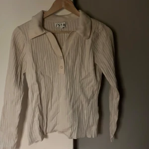 Beige ribbad tröja från Ista - Säljer en stilren beige ribbad tröja från Ista med krage och knappar framtill. Tröjan har långa ärmar och en klassisk design som passar perfekt för både vardag och mer uppklädda tillfällen.