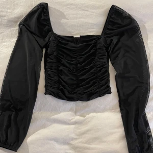 Svart långärmad topp - Snygg svart topp med långa ärmar och rynkade detaljer framtill. Toppen har en fyrkantig urringning och är croppad för en trendig look. Perfekt för att styla med jeans eller kjol.