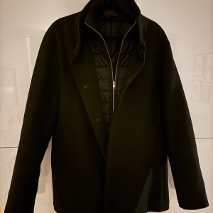 Svart kappa från Jack & Jones - Snygg svart kappa från Jack & Jones i storlek M. Den har en stilren design med dragkedja och knappar framtill. Perfekt för kyligare dagar med sin quiltade insida som håller värmen. Ett måste för garderoben!