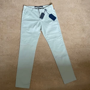 Ljusbeige chinos från AT.P.CO - Snygga vita/ljusbeige chinos från AT.P.CO i slim fit. Byxorna har en stilren design med knappar och dragkedja. Perfekta för en elegant look. Tillverkade i ett bekvämt material med elastan för extra stretch.