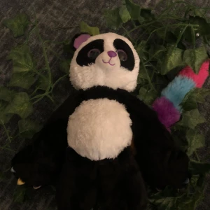 Panda gosedjur med regnbågsfärgad svans - Mjuk och gosig panda med en unik regnbågsfärgad svans. Perfekt för kramar och lek. Pandan har en klassisk svart och vit design med stora, vänliga ögon.