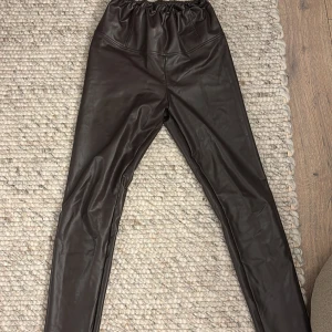 Bruna skinnleggings från Sanne Alexandra - Snygga bruna leggings i skinnimitation från Sanne Alexandra. De har en elastisk midja för extra komfort och en tight passform som ger en stilren look. Perfekta för att höja din outfit till nästa nivå.