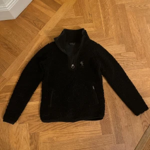 Svart fleece tröja från Kingsland - Mysig svart fleece tröja från KL med halv dragkedja och hög krage. Tröjan har långa ärmar och två praktiska fickor med dragkedja framtill.