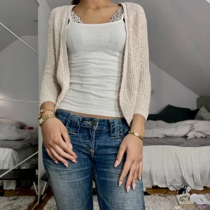 Beige stickad kofta - Säljer en stilren beige stickad kofta med långärmad design. Perfekt för att lägga till ett lager värme och stil till din outfit. Koftan är öppen framtill och har en mjuk och bekväm passform.