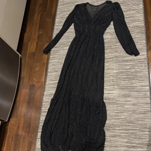 Svart glittrig långklänning - Elegant svart långklänning med glittriga detaljer och v-ringning. Klänningen har långa ärmar och en smickrande passform som ger en stilren look. Perfekt för speciella tillfällen.