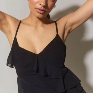 Svart långklänning med volangdetaljer - Elegant svart långklänning med smala axelband och volangdetaljer längs sidan. Klänningen har en slits som ger en stilfull touch. Perfekt för en kväll ute eller en speciell tillställning. Köpt för 600kr! Använd en dag, som ny!!🥰 