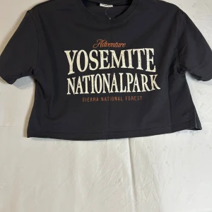 Svart croppad t-shirt från Kappahl - Säljer en svart croppad t-shirt från Kappahl med tryck 'Yosemite National Park' i vitt och orange. Perfekt för en avslappnad stil. T-shirten har korta ärmar och rund halsringning.