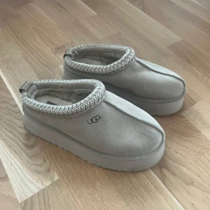 Beige mocka tofflor från UGG - Säljer ett par bekväma beige tofflor från UGG i mocka med mjuk insida. De har en robust sula och dekorativ stickad kant runt öppningen. Perfekta för att hålla fötterna varma och sköna. storlek 37