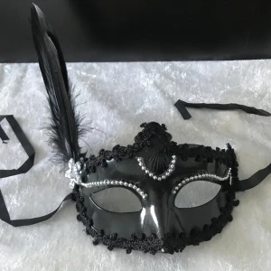 Svart maskeradmask med fjäder - Elegant svart maskeradmask med dekorativa pärlor och en stor fjäder på sidan. Masken har ett glänsande yttre och knyts med band. Perfekt för maskerad eller festliga tillfällen.  Helt ny, finns vit och svart färg kvar 