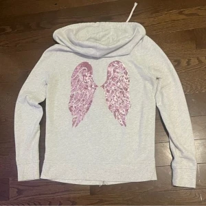Rare victoria secret zip hoodie med vingar - Säljer en snygg rare grå hoodie från Victoria's Secret med rosa paljettvingar på ryggen. Tröjan har dragkedja och en mysig huva. Perfekt för en avslappnad stil med en touch av glamour. Har använt minst 2 gånger och inga hål eller fläckar. 
