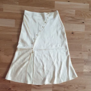 Krämfärgad långkjol från NA-KD - Elegant krämfärgad långkjol från Mathilde Gøhler x NA-KD. Kjolen har en omlott-design med dekorativa knappar och en subtil slits framtill. Tillverkad i ett mjukt och glansigt material som ger en lyxig känsla.