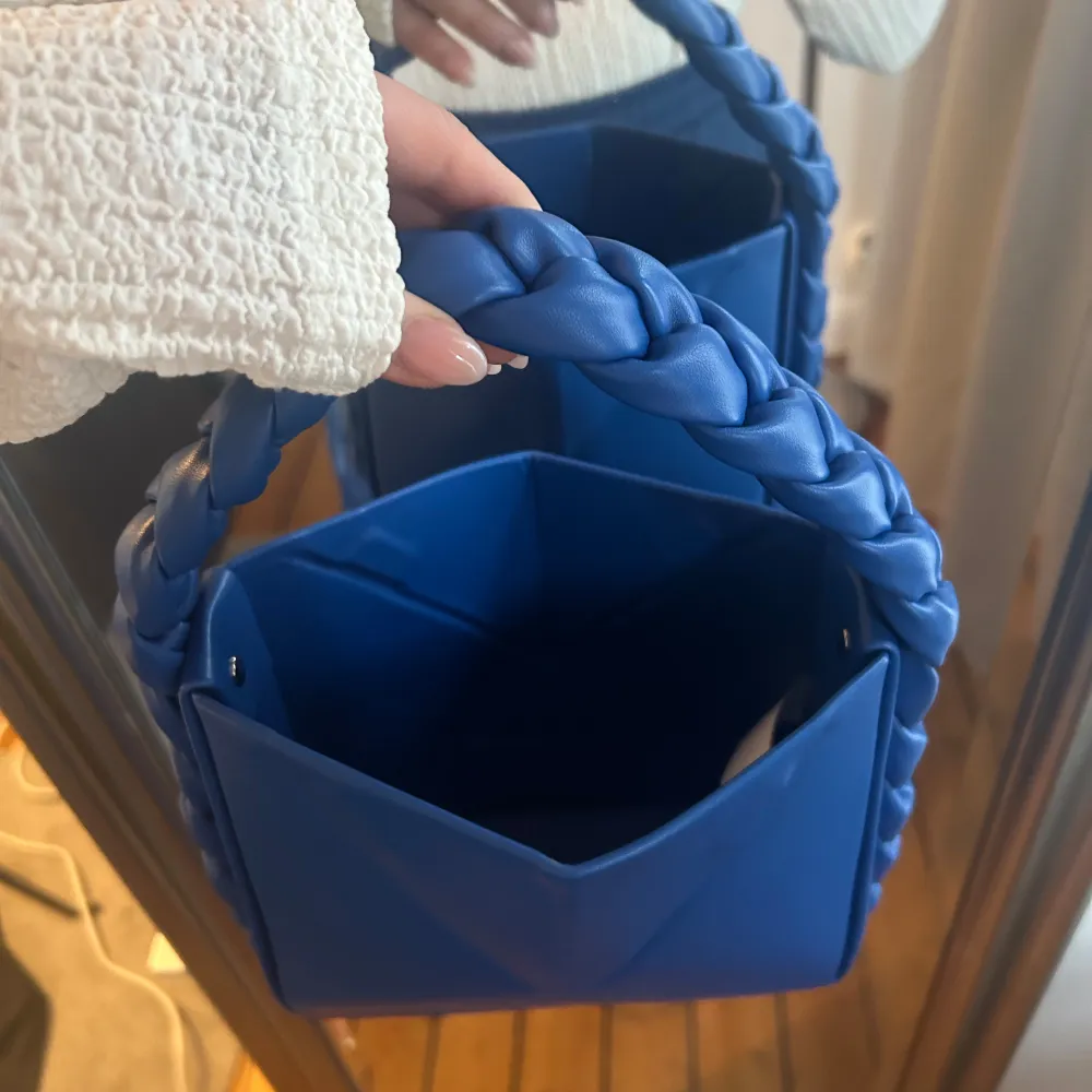 Snygg blå axelväska med ett unikt flätat handtag. Väskan har en modern design med en triangulär form och är tillverkad i syntetmaterial. Perfekt för att ge din outfit en färgklick. Har magnet vi öppning vilket gör att man kan forma den på olika sätt.. Laukut & Käsilaukut.