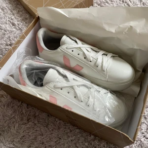 Vita sneakers från Veja med rosa detaljer - Snygga vita sneakers från Veja med rosa detaljer. Skorna har en klassisk design med snörning och en bekväm sula. Perfekta för en stilren look.