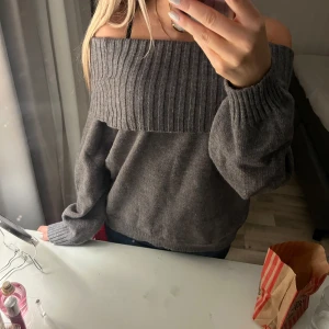 Grå offshoulder tröja stickad  - Säljer en snygg grå offshoulder tröja med ribbad detalj upptill. Tröjan har långa ärmar och en loose passform, perfekt för en avslappnad stil. Den är gjord i ett mjukt material som känns skönt mot huden. Oanvänd